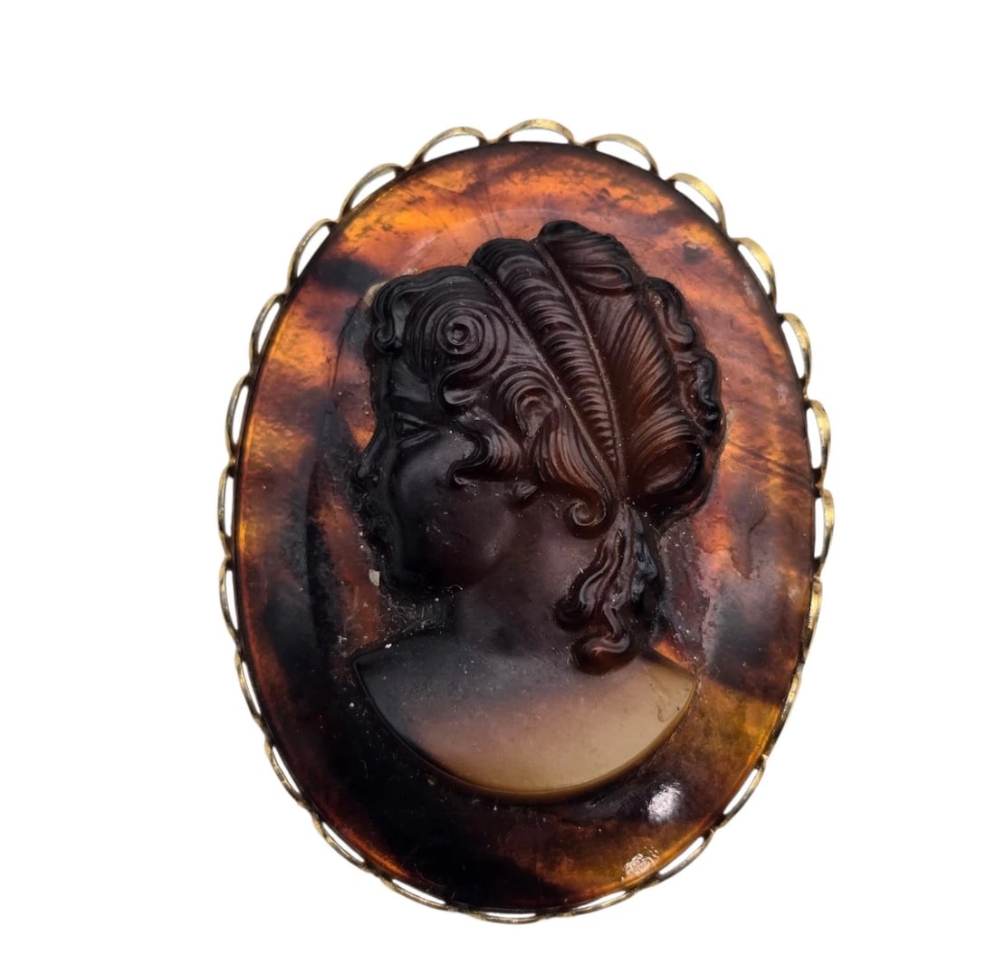 Vintage Amber Brown Cameo Brooch Oval Gold Tone Background: oval vintage brooch featuring a high-relief cameo portrait of a woman's profile with detailed, styled hair. The piece is crafted from a glossy, semi-translucent faux-tortoiseshell material with amber 