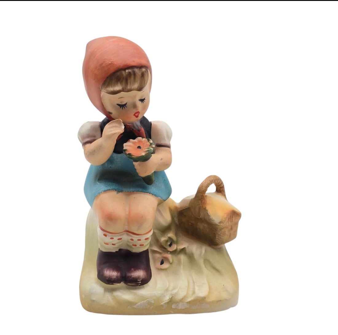 VINTAGE 1950'S ERICH STAUFFER ARNART CREATIONS PICNIC TIME CROWN SYMBOL JAPAN: A lovely vintage authentic Goebel Hummel porcelain figurine featuring a young girl seated on a grassy knoll. She wears a prominent red headscarf and a blue dress, gently looking down at a blooming pin