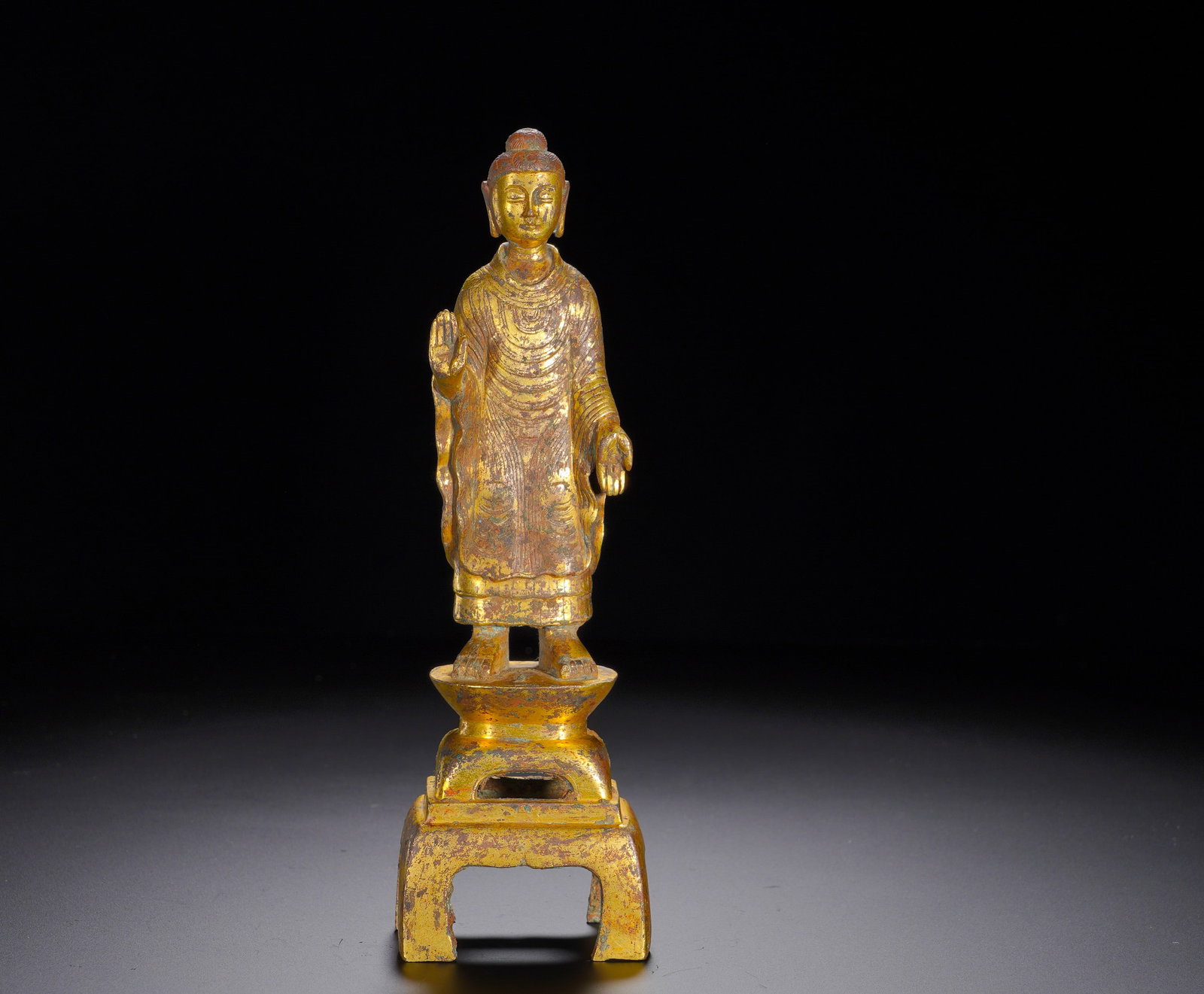 A gilded bronze standing statue of Sakyamuni Buddha from the Northern Dynasties: Height:21cm(8.27in), Width:6.5cm(2.56in), Weight:660g (1.455lb)His right hand forms Abhaya Mudra to grant fearlessness, while the left hand extends in Varada Mudra to bestow blessings. The surviving g