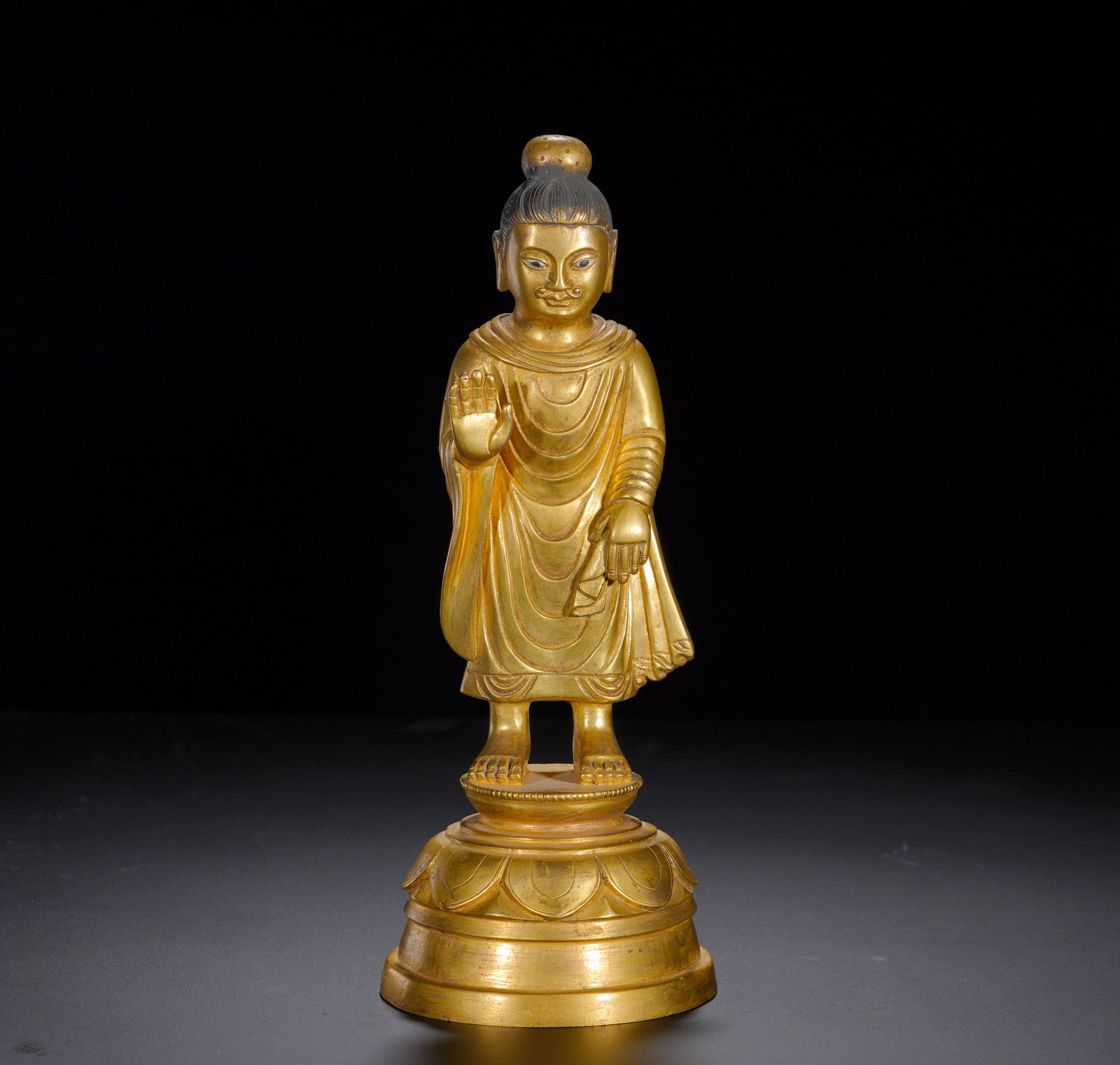 A gilded bronze standing Buddha of Sakyamuni from the Qing Dynasty: Height:25cm(9.84in), Width:9.5cm(3.74in), Weight:1170g (2.579lb)He stands barefoot on a two-tier circular lotus base with full petals decorated by a band of beading. Integrating Mathuran artistic heri