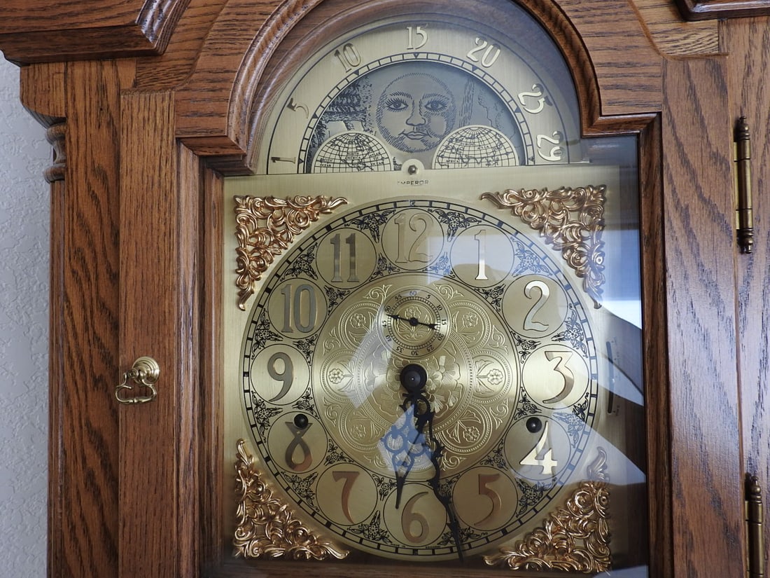 Emperor Oak & Brass Grandfather Clock: Emperor grandfather clock with solid oak case, brass-tone dial and pendulum, German-made movement per notes, original storage key, and original winding key. Approx. 87 x 23 x 14 in.