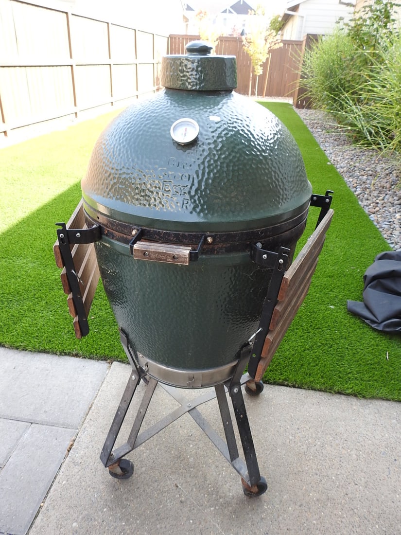 Big Green Egg Ceramic Kamado Smoker Grill with Cart and Cover: Big Green Egg ceramic kamado smoker grill with rolling metal cart, side shelves, thermometer, top vent, and fitted cover. Approx. 48 x 28 x 28 in.