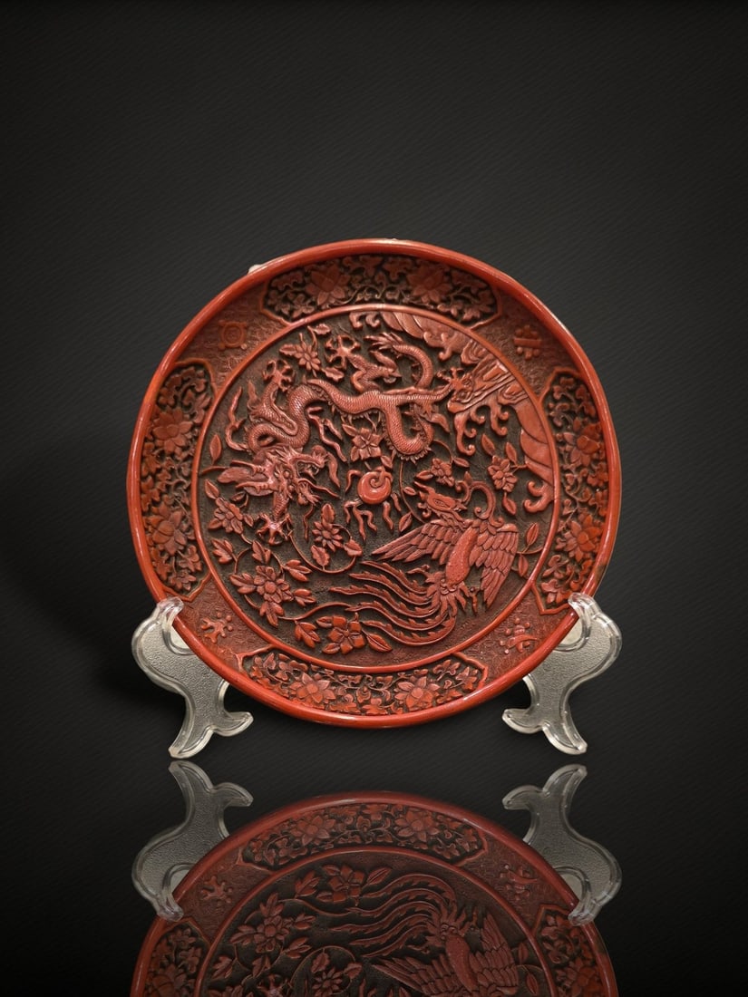 A Chinese Carved Cinnabar Lacquer 'Dragon and Phoenix' Dish: A carved cinnabar lacquer dish decorated in relief with a five-clawed dragon and a phoenix confronting a central flaming pearl, amid dense floral scrolls and foliage reserved on incised diaper grounds