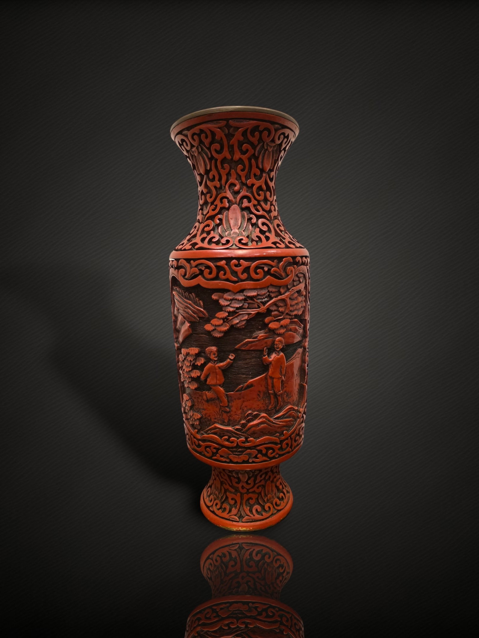 A Chinese Carved Cinnabar Lacquer 'Figures in a Landscape' Vase: A baluster vase decorated in carved red lacquer of cinnabar type over a metal (apparently brass or bronze) body, with a slightly everted mouth, waisted neck and spreading foot. The principal register 