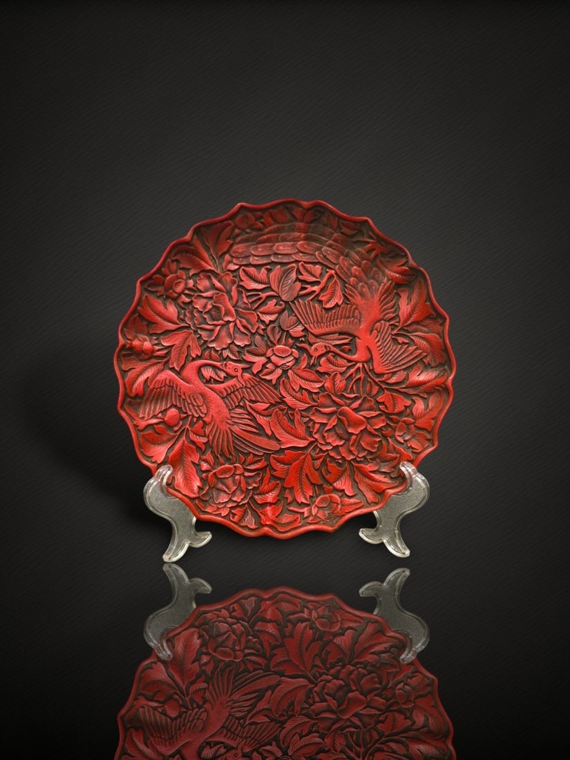 A Chinese Carved Cinnabar Lacquer 'Phoenix and Peony' Dish: A shallow dish with a barbed, foliate rim, worked in the manner of carved cinnabar lacquer over a darker ground. The interior is deeply and densely carved with two long-tailed phoenixes in flight amon