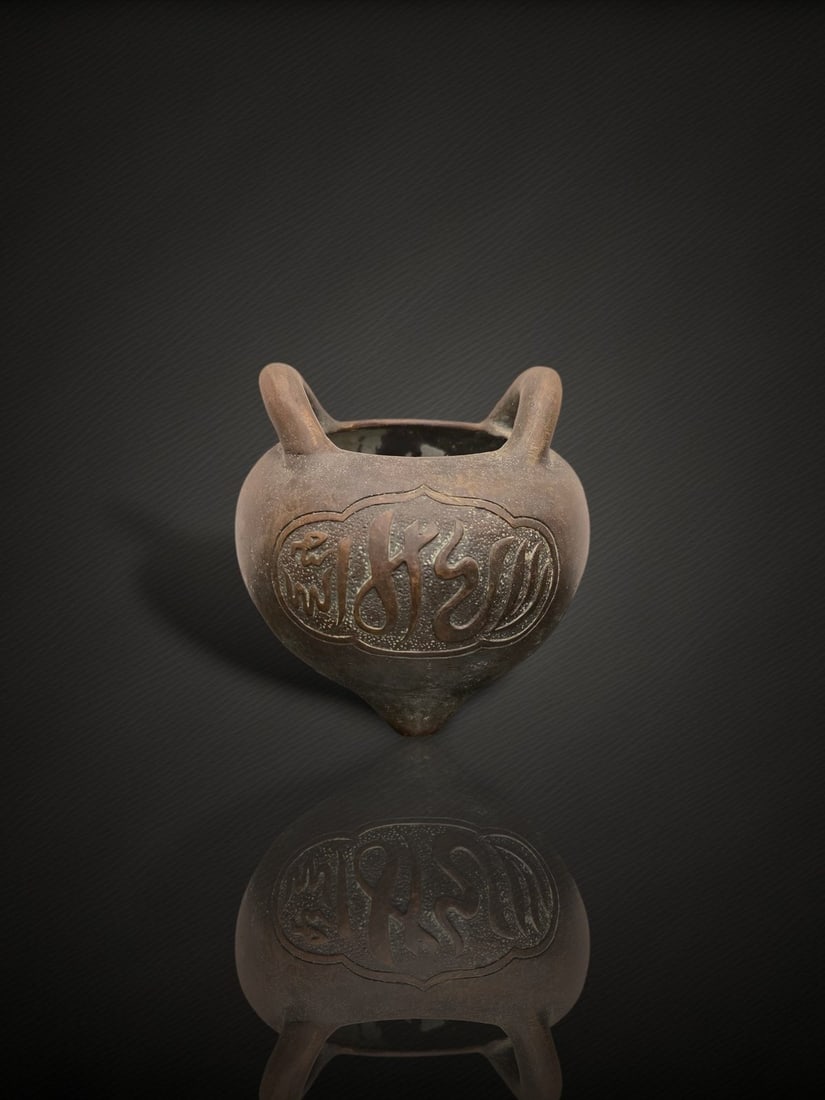 A Chinese Bronze 'Arabic Inscription' Tripod Censer: A cast bronze incense burner of rounded, slightly pear-shaped form raised on three feet, with a pair of upright loop handles set on the everted rim. The body is decorated in relief with a lobed cartou