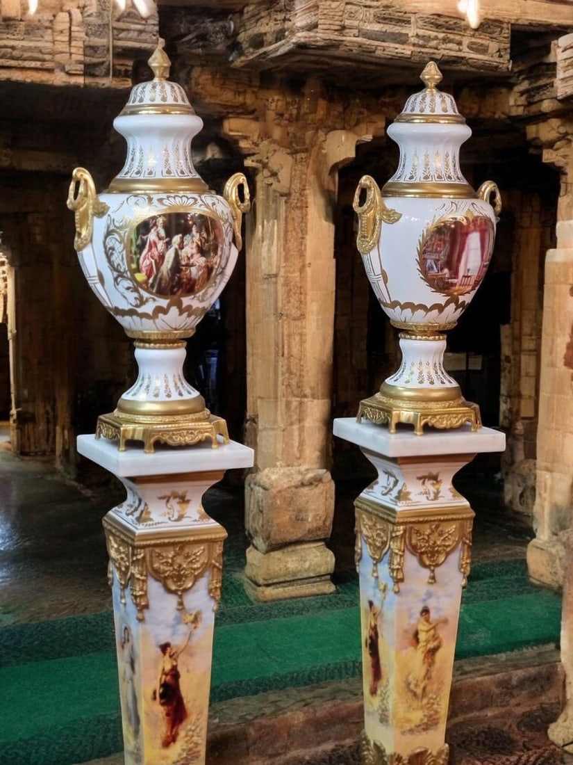Antique-Style Porcelain Urn Set on Pedestals –: Description: This exquisite pair of urns sits atop intricately decorated porcelain pedestals, each crafted with meticulous detail and ornate artistry. The urns feature classical scenes - one showing a