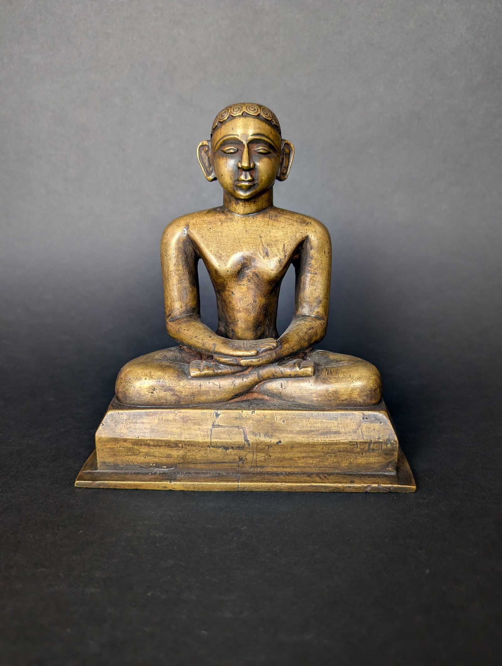 Jain Tirthankara possibly Sambhavanatha. Heavy brass casting, 20th c: The meditating Tirthankara is not readily identifiable. There are small dots under his feet which seem to portray a horse jumping behind a tree. The horse is the insignia for the 3rd tirthankara, Samb