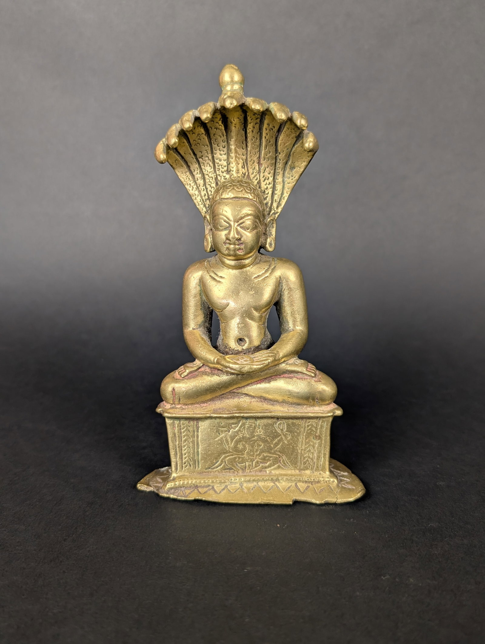 Parsvanatha seated under 9 headed cobra: Parsvanatha sits under the canopy of a 9 headed cobra, protecting his meditation. He is wearing earrings, or perhaps it is just an extended earlobe? In addition, his navel is rather large. On his thro