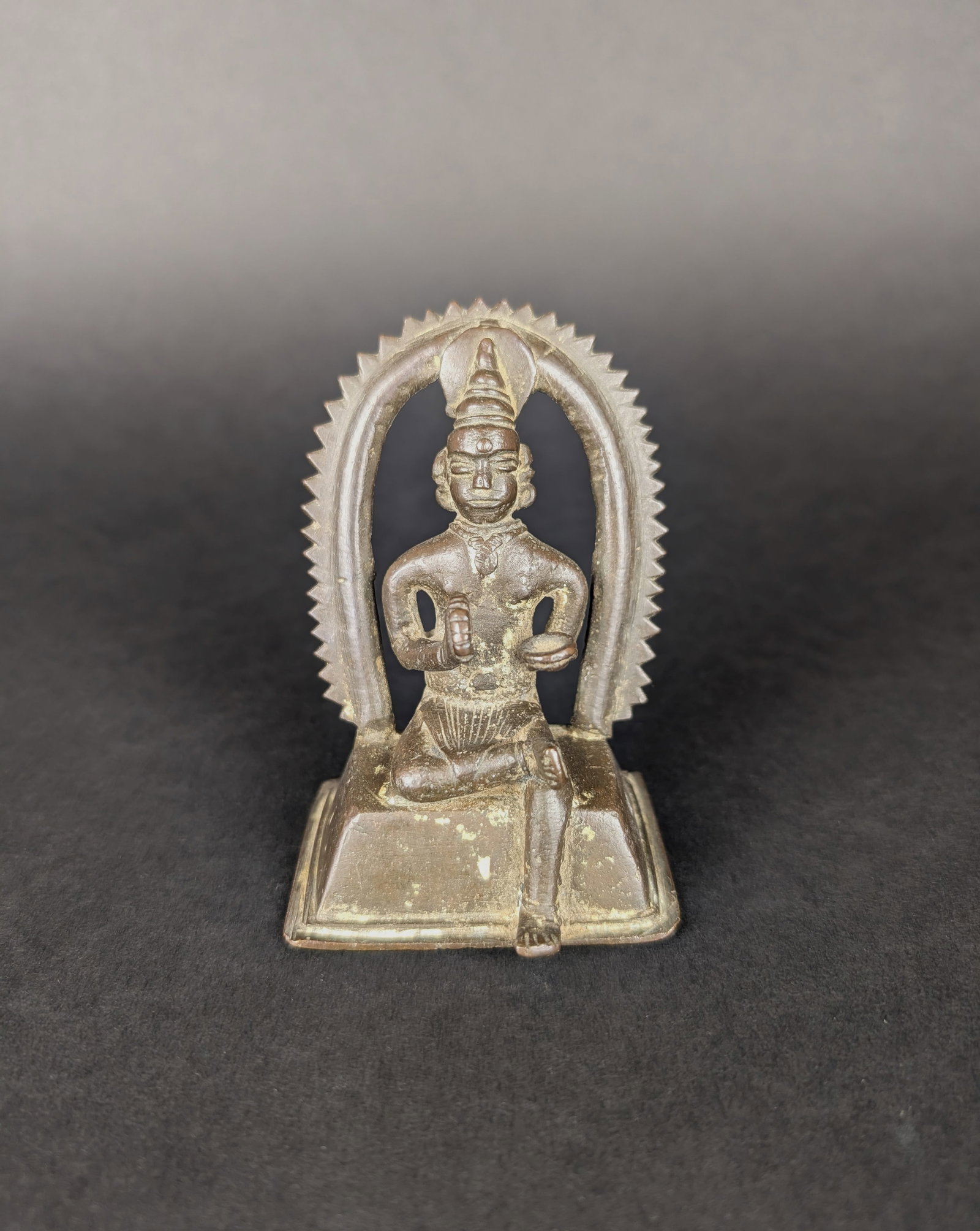Seated Ascetic, Folk-Style: The seated Ascetic is a Hindu ascetic from Kerala. The prabhavali and the halo behind his head suggest a divinity. He is holding prayer beads and a container. His crown is elaborate for the