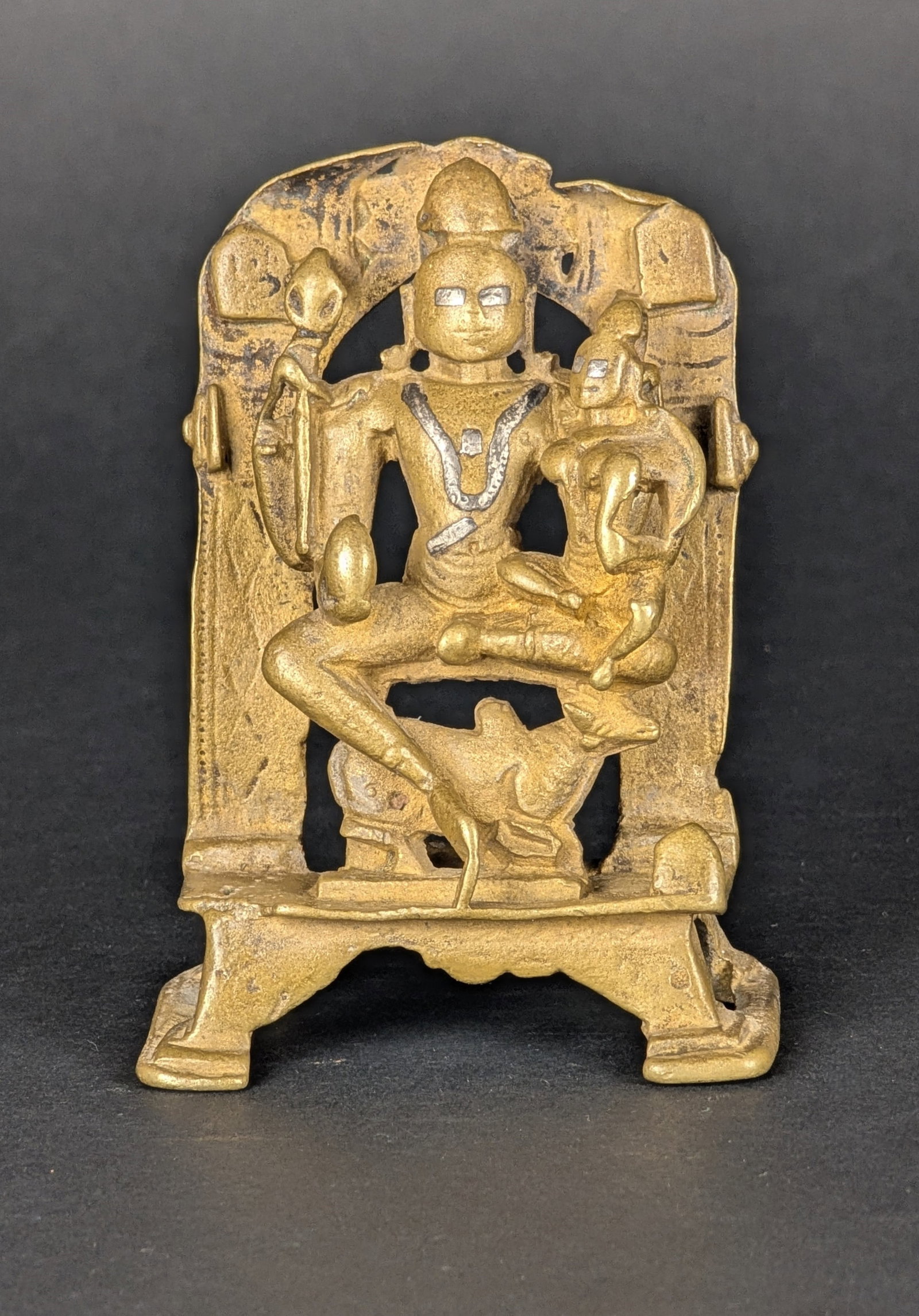 Uma Maheshvara, pre-16th c: This bronze of Umamaheshwara, Shiva and Parvati is aged. In his rear hands, Shiva is holding Trishul and a cobra, an unusual and rare combination. His right lower hand is holding a fruit. On his head 