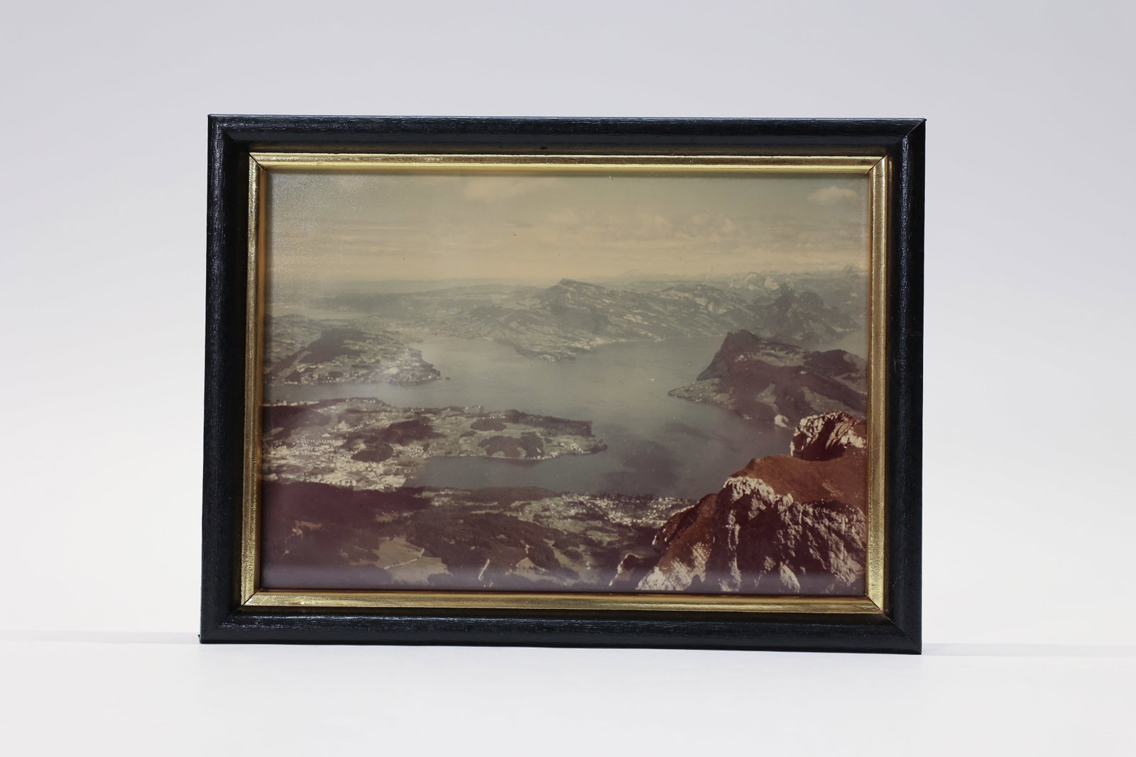 Vintage Framed Photo, European Mountain Lake: A serene vintage color photograph captures a sweeping elevated view over a still alpine lake, framed by dramatic snow-dusted peaks and terraced hillside villages below. The muted, atmospheric palette 