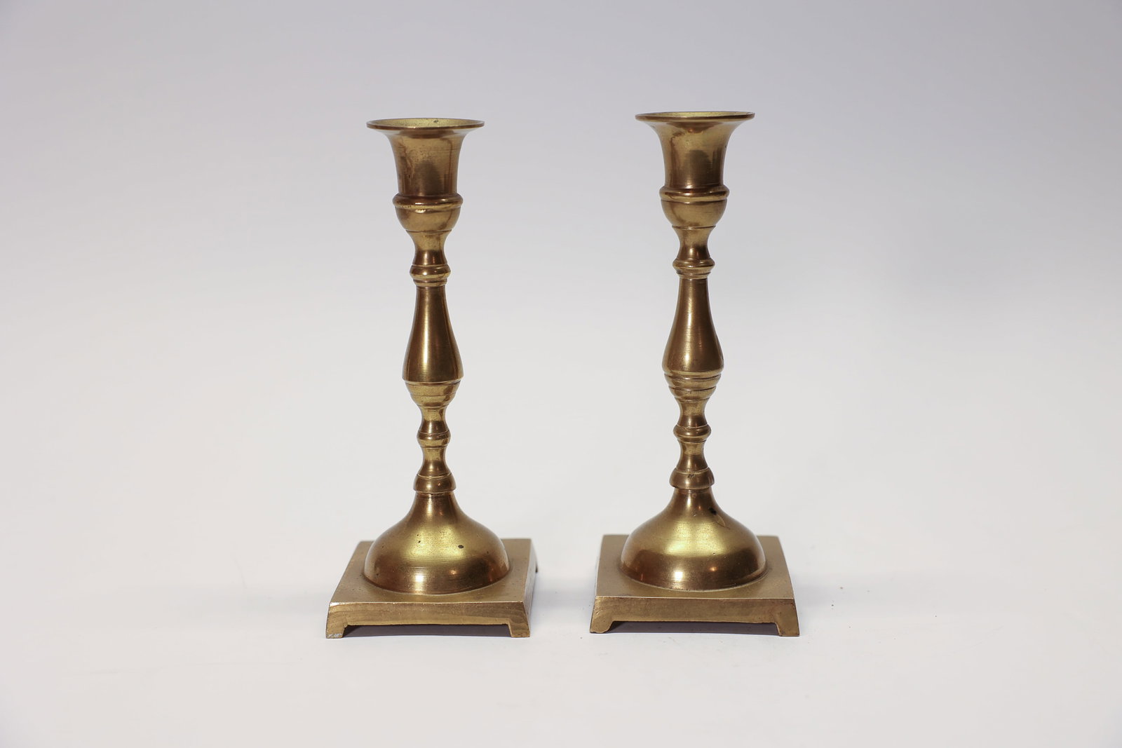 Pair Brass Candlesticks India 6 1/4" Tall: A handsome pair of solid brass candlesticks from India, each standing 6 1/4 inches tall on a square footed base. The classic turned baluster stem features a bell-shaped bobeche, ring-turned detailing 