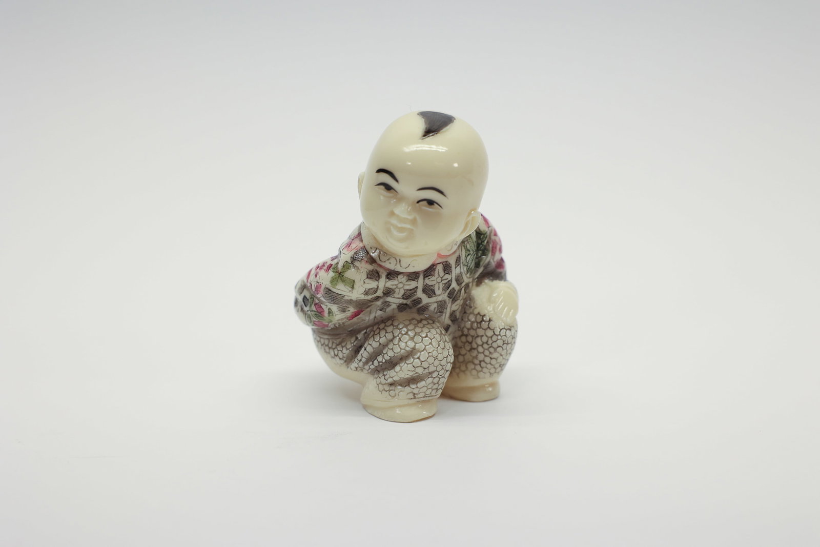 Carved Figural Squatting Child in Floral Robe: Carved figural okimono-style piece depicting a squatting bald child in a floral and lattice-patterned robe, one hand resting on a rounded object (peach or gourd). Detailed facial features with