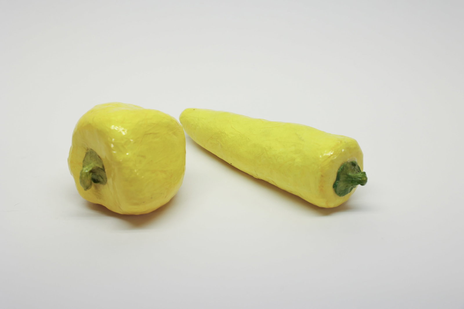 Pair of Lacquered Papier-Mâché Yellow Peppers: Decorative faux vegetables in lacquered papier-mâché, modeled as a yellow bell pepper and a yellow hot pepper, each with a hand-formed green stem. Glossy finish throughout.Dimensions: H: 3", W: 5",
