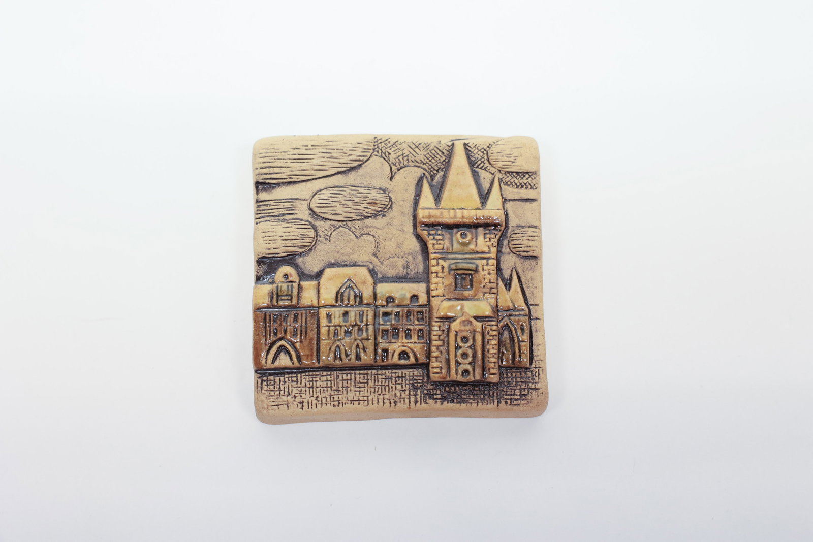 Signed Studio Pottery Tile, European Townscape: Hand-carved stoneware tile depicting a European old-town skyline, with a tall Gothic tower featuring twin spires and stepped brickwork rising above a row of gabled townhouses. The scene is rendered in