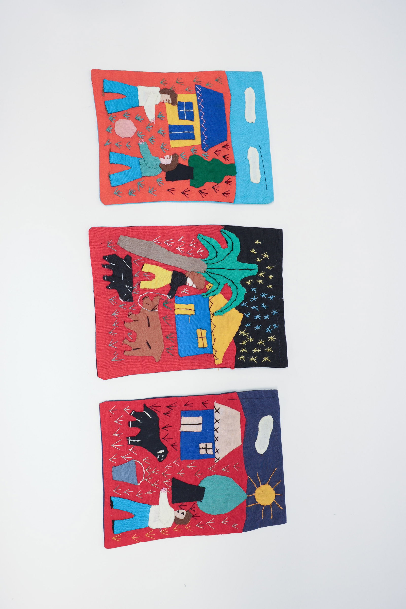 Set of 3 Colombian Appliqué Folk Textiles: Set of three hand-appliquéd cotton folk art textiles, made in Colombia, each depicting a colorful rural village scene with figures, houses, animals, and trees built up in layered fabric shapes and ha