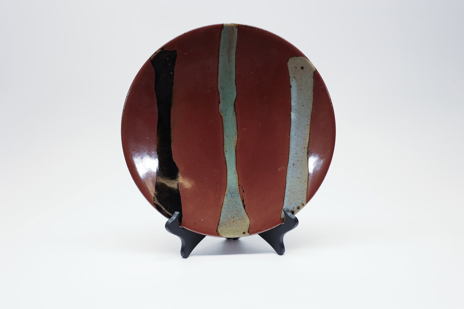 Studio Pottery Charger, Iron Red Glaze: Hand-thrown studio pottery charger with a deep rust-red glazed surface, accented by three vertical glaze bands in black, celadon-green, and cream running from rim to rim. The reverse shows a wide, ung