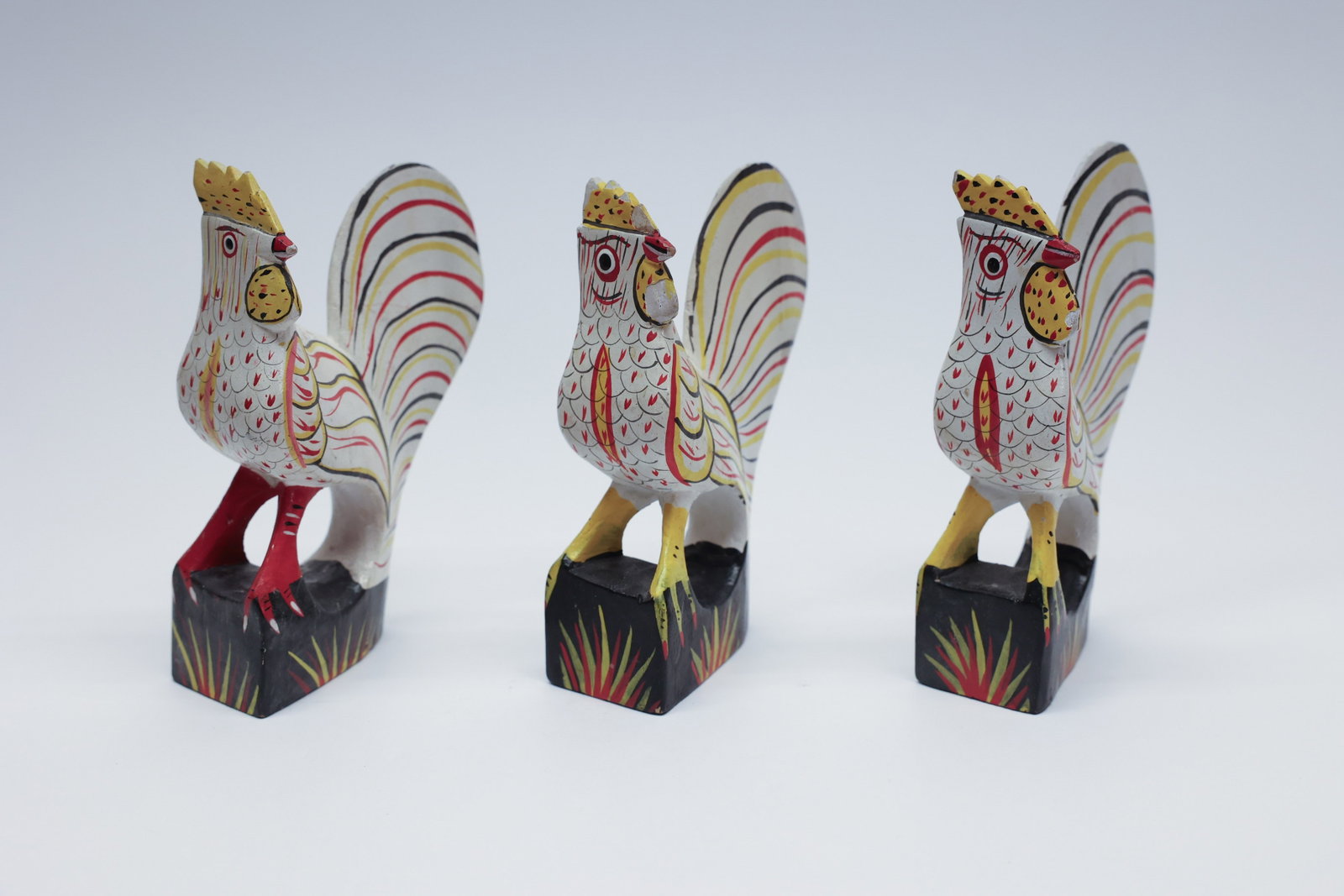 Set of 3 Carved Folk Art Painted Roosters: Set of three hand-carved and painted wooden roosters, each perched on a black wedge-shaped base decorated with stylized foliage. The roosters feature bold, graphic feather patterning in red, yellow, a