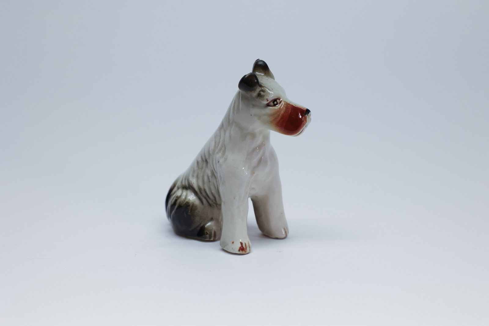 Vintage Porcelain Wire Fox Terrier Figurine: This porcelain figurine depicts a wire-haired fox terrier seated upright and alert, its rough coat texture suggested through fine incised lines across the back and haunches, with crisp black-tipped ea