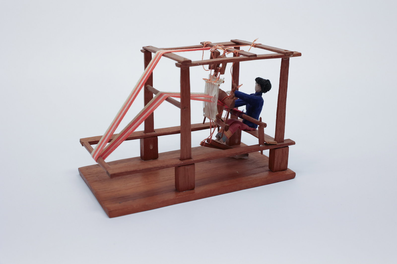 Miniature Wood Loom Weaver Diorama: Description:A charming handmade folk-art diorama depicting a weaver at work on a wooden frame loom, with a small cloth-bodied figure dressed in a blue jacket and striped skirt seated at the loom's ben