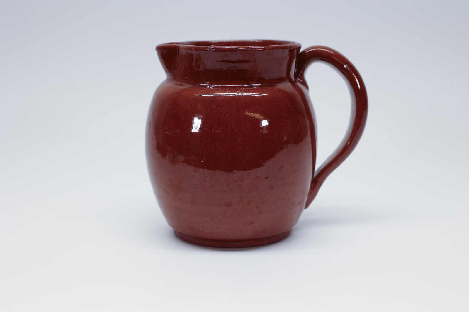 Vintage Spanish Brown Glaze Pottery Pitcher: Small studio-style pottery pitcher with a bulbous, rounded body, pulled loop handle, and pinched spout, finished in a glossy reddish-brown glaze that lightens to a mottled, warmer tone in spots where 