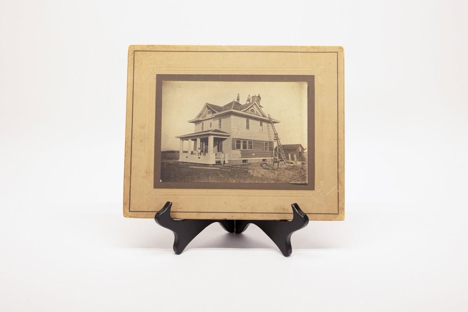 Antique Cabinet Photo Victorian House Moving Build: Description:Large-format sepia-toned photograph, mounted on its original decorative embossed paper board, showing a two-story Victorian/Queen Anne style house raised on wooden rails or beams, with