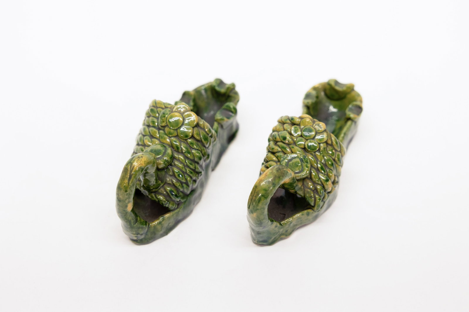 Pair Green Majolica Pinecone Ashtray Slippers: Pair of glazed ceramic ashtrays modeled as curled-toe slippers, each with a raised loop handle at the toe and a textured, bumpy pinecone/scale surface covering the upper portion, finished in a