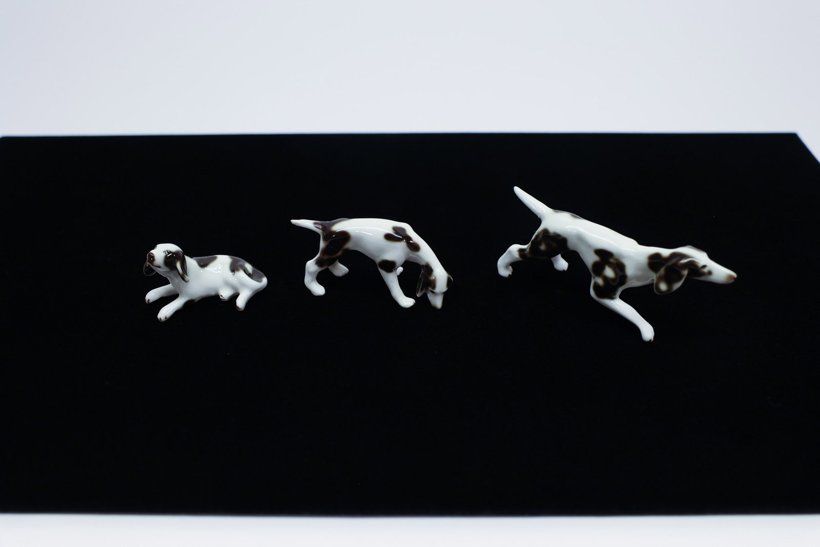 3 Miniature Porcelain Spaniel/Setter Figurines: Description:Set of three miniature porcelain sporting dog figurines, likely spaniels or pointers, each glazed white with brown liver-colored patches, captured in three different dynamic poses — lyin