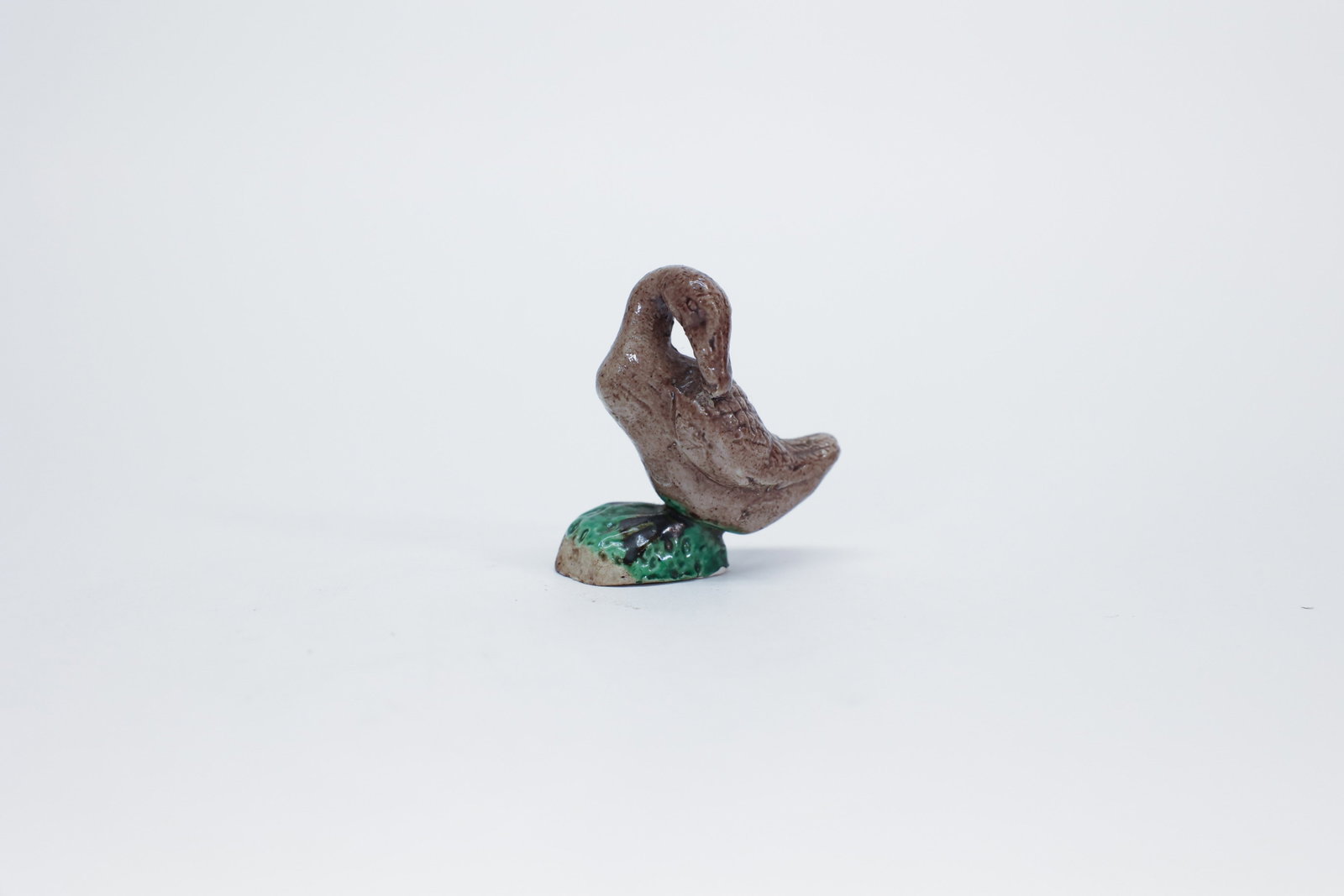 Mini Chinese Mud Duck/Goose Ceramic Figurine: Description:Small hand-formed clay figurine of a goose or duck in a preening pose, head curved down toward its back, glazed in mottled tan-brown tones with incised feather detailing, standing atop a