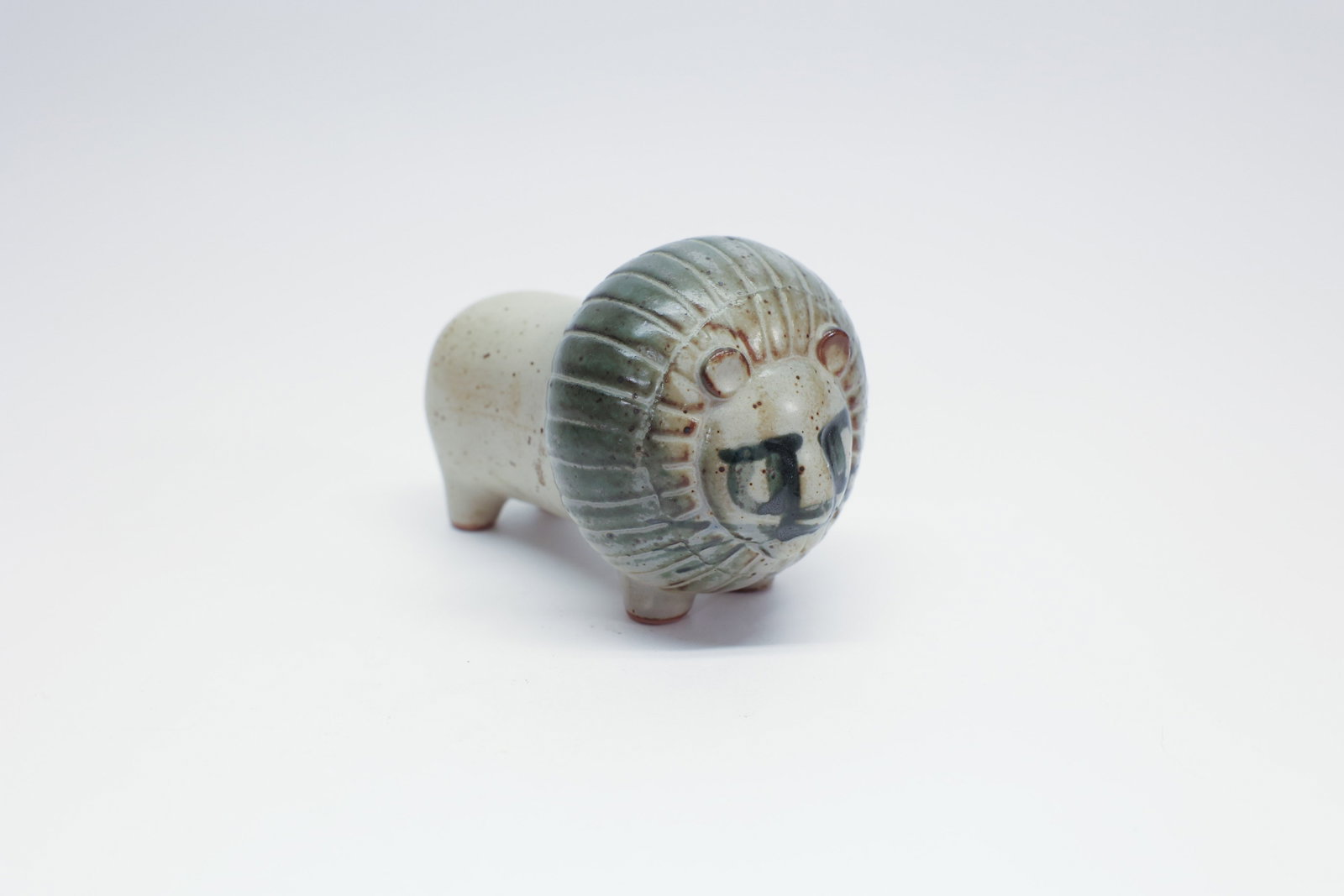 Mid Century Scandinavian Ceramic Lion Figure Japan: Description:A charming, modernist ceramic lion figurine with a stylized, cartoon-like form: a rounded body raised on four short legs, a large disc-shaped head, and a ribbed mane rendered in a sage-gre
