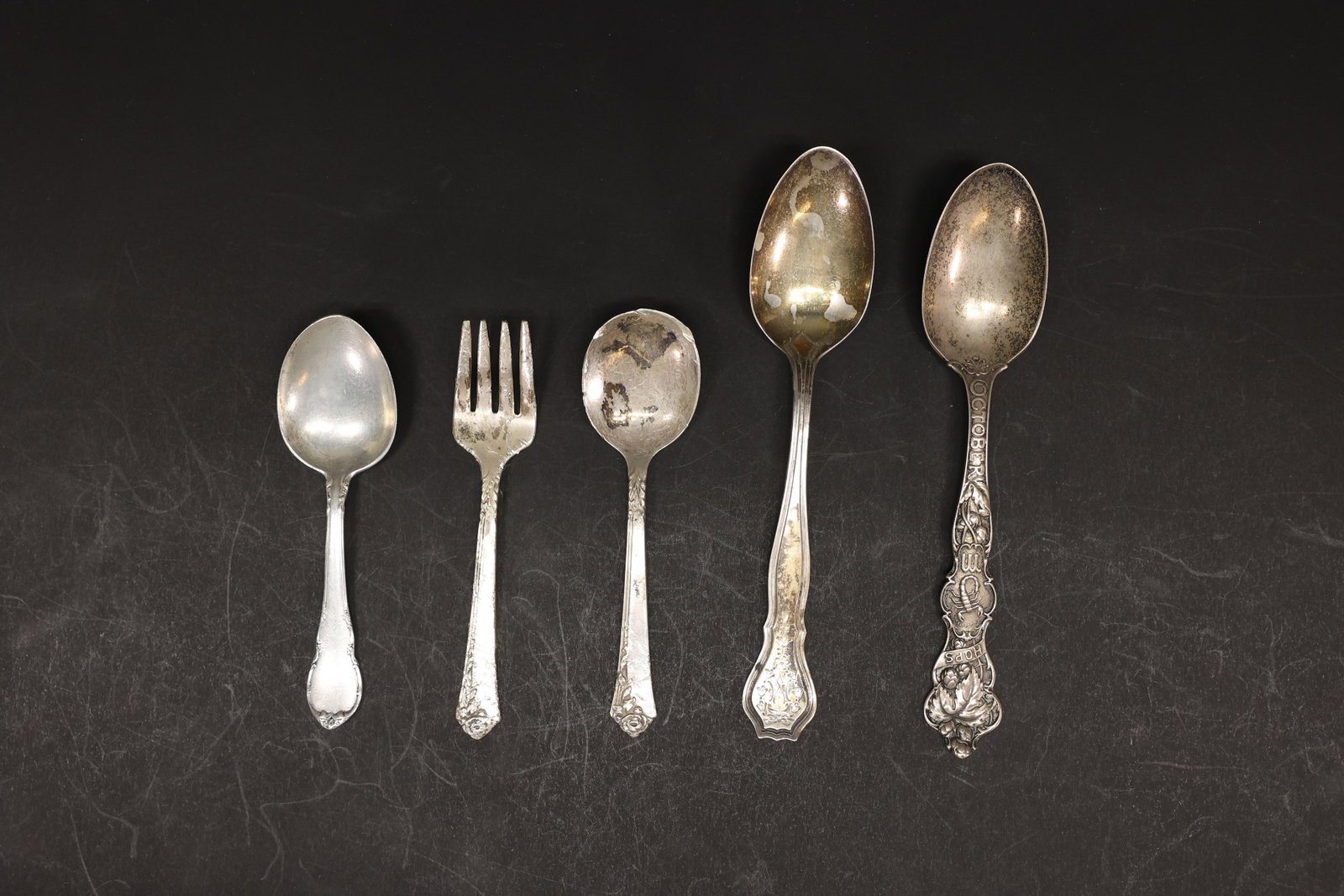 Mixed Sterling Silver Flatware Lot, 5 Pieces: Group of five sterling silver flatware pieces from different patterns and makers: a teaspoon, a fork, a small spoon, a large serving spoon, and an October-themed souvenir spoon with a figural handle.