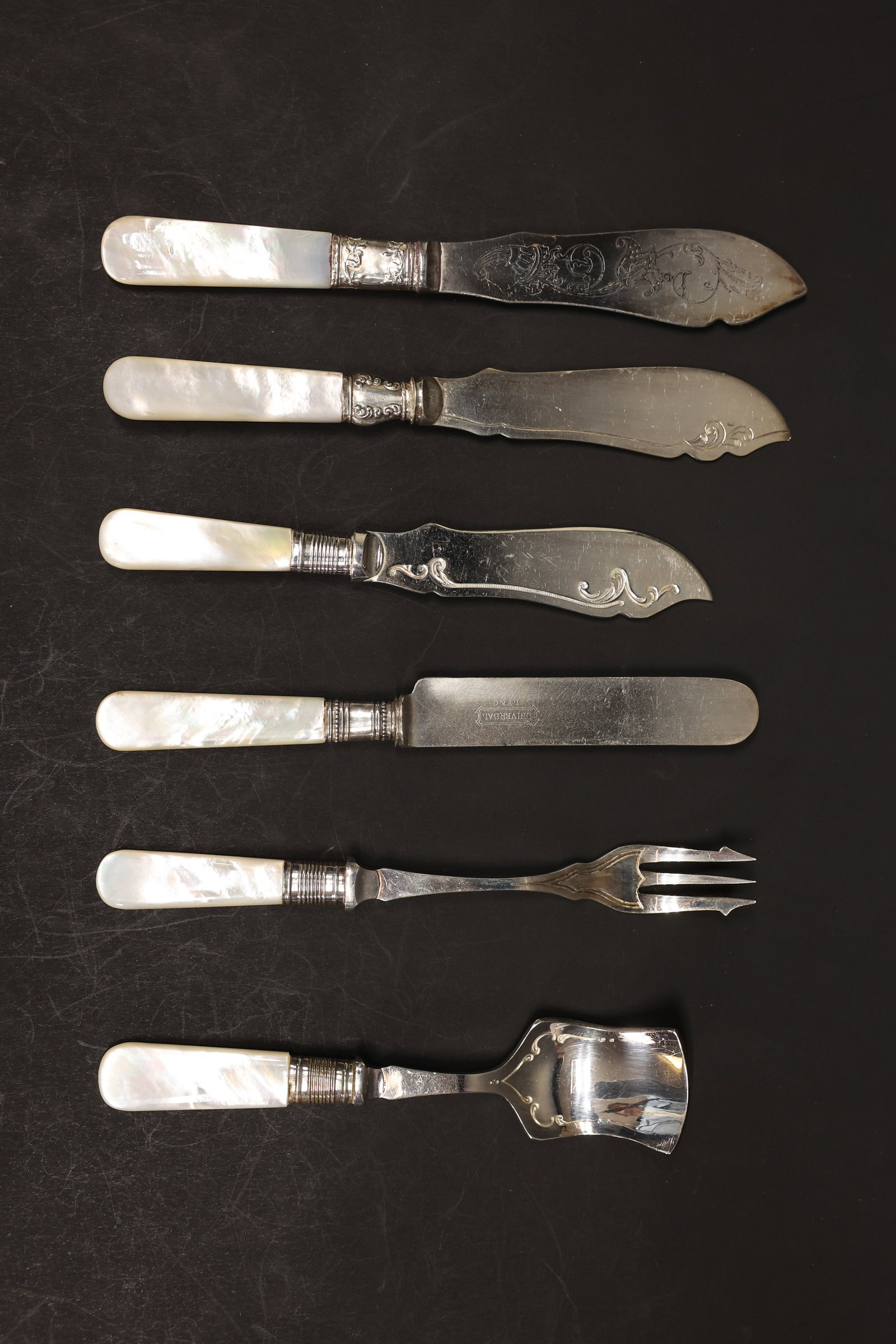 Mixed Mother-of-Pearl Handle Flatware, Set of 6: Group of six mother-of-pearl handled serving pieces with silver mounts: four flat-bladed fish/butter knives with engraved and scroll-etched blades, a three-tine pickle/olive fork, and a wide flat-blad