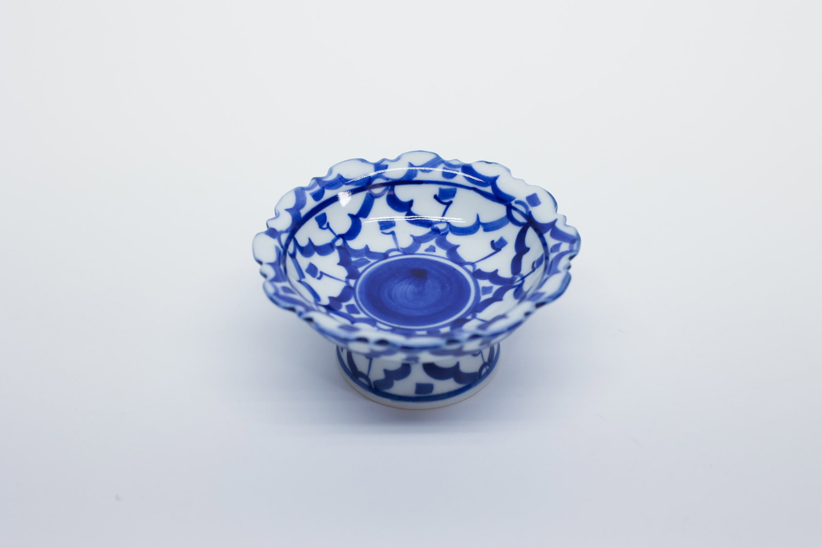 Thai Blue & White Porcelain Footed Salt: Small footed porcelain dish with a scalloped, fluted rim, hand-painted in underglaze blue with a dense floral/scroll motif and a solid dark blue medallion at the center of the well. Pedestal foot deco