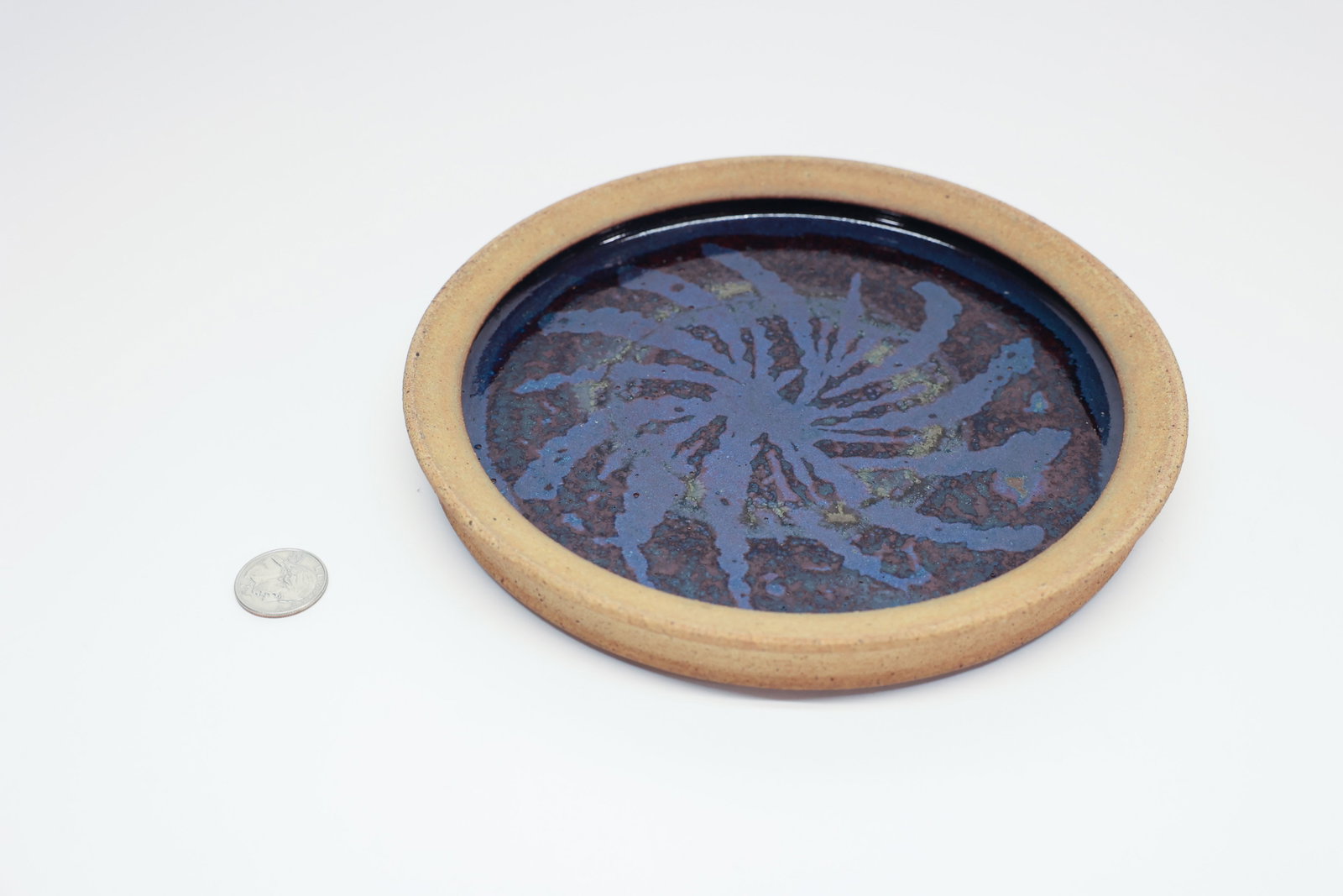 Oscar Bucher Stoneware Plate, Crater/Lava Glaze: Hand-thrown stoneware plate with a wide unglazed buff rim and a deep blue crater/lava glaze pooled in the well, showing a radiating crackled texture with flashes of green and rust. Underside inscribed
