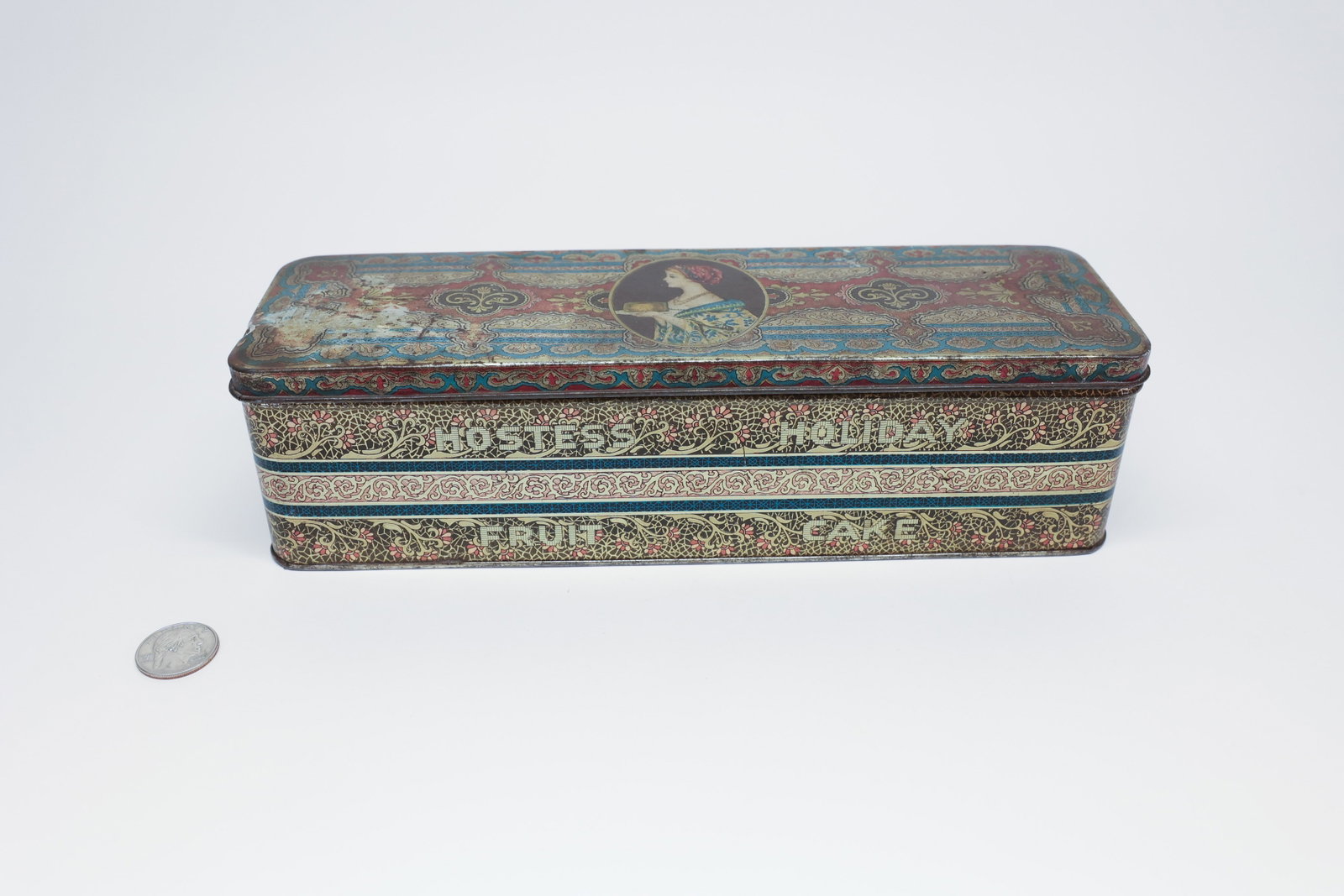 Metal Package Corp. Hostess Fruit Cake Tin: Lithographed tin box, rectangular with rounded corners, in a Victorian-revival pattern of red, teal, and gold scrollwork bands. Center medallion on lid depicts a woman in profile holding a covered cak