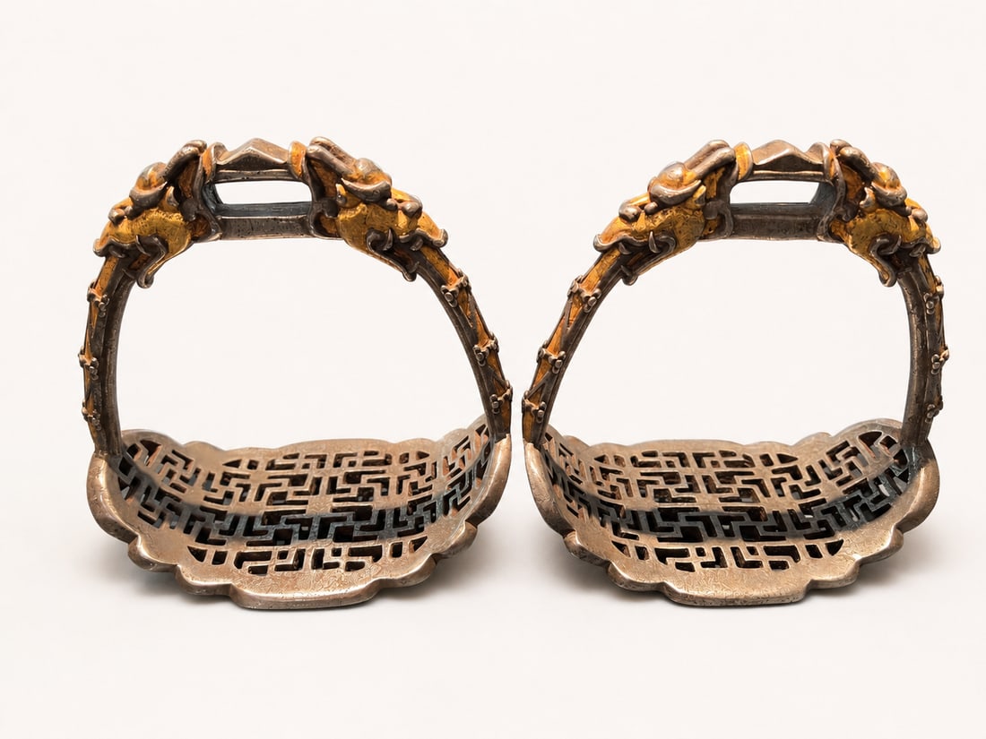 A Pair of Gold-Damascened Iron Stirrups with Dragon Heads, Qing Dynasty: A pair of iron stirrups with gold decoration, attributed to the Qing dynasty. Each stirrup has an arched frame rising to a rectangular strap aperture, flanked by sculptural dragon heads with pronounce