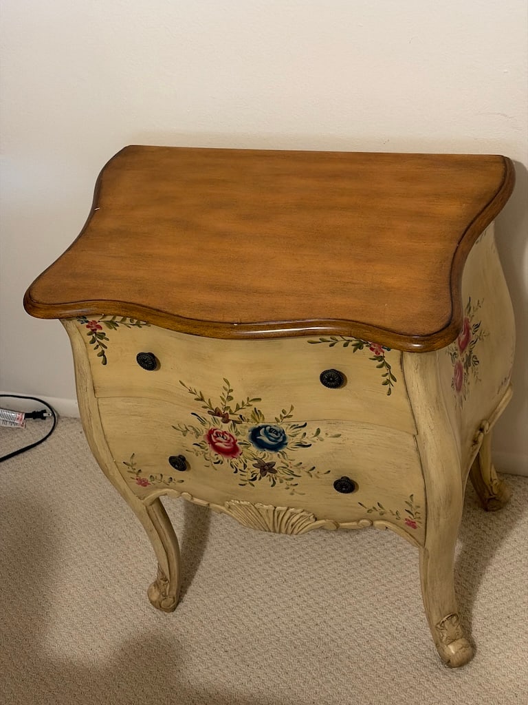 Coastal Cottage Painted Wood Coffee Table with Drawer: A compact painted wood bombe-form chest with a warm brown serpentine top, cream-colored case, floral rose decoration, dark round drawer pulls, carved-style apron, and cabriole legs. A rear label reads