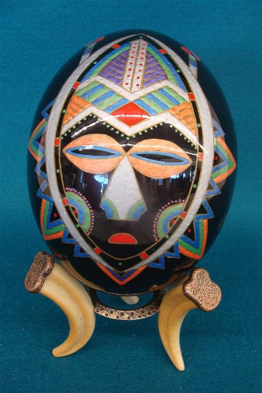 Hand painted Ostrich Egg a South African Knife Sheath M: Hand painted Ostrich Egg a South African Knife Sheath Mask from the Baule Atutu Tribe. Artist Signed Mazo. Comes with Carved Faux Boar Tusk Stand. Auction Estimate: $50-100.