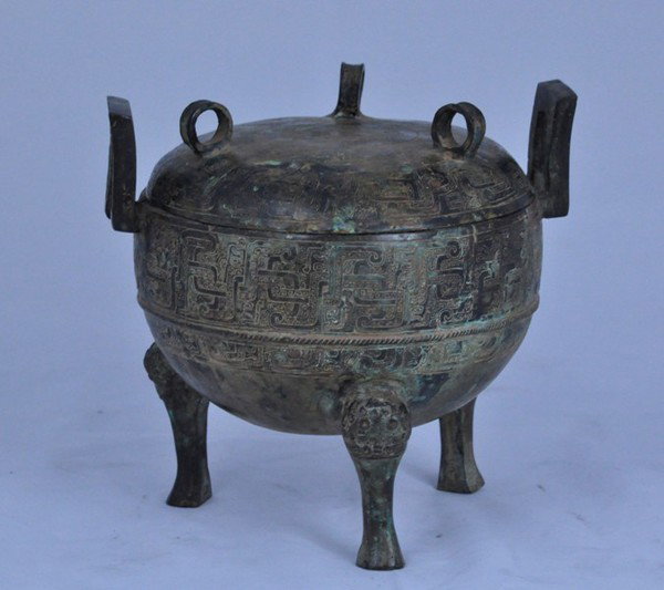 ARCHAIC BRONZE RITUAL TRIPOD FOOD VESSEL & COVER: the globular body cast with a thin band of hooked scrolls and chevrons, set with two upright arch handles with addorsed dragons and rope-twist borders, the flat rounded cover with a further scroll-and
