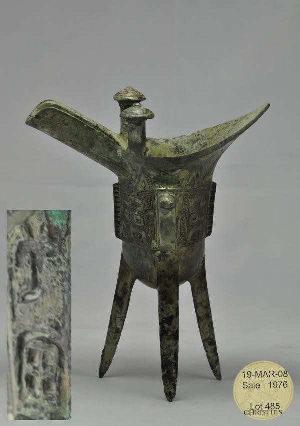 A BRONZE TRIPOD RITUAL WINE VESSEL, JUE (SHANG): Raised on three blade-form supports, the sides flat-cast with a band comprising two taotie masks with rounded rectangular eyes centered on and separated by notched flanges, below a band of blades, wit