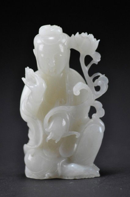 A WHITE JADE CARVING OF A GUANYIN.: The immortal shown standing holding a peony stem bearing two blossoms, wearing long layered robes falling in graceful folds and a scarf draped around the back and over the arms, the hair drawn up into