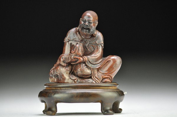 A SOAPSTONE LUOHAN AND LION CUB GROUP.(QING DYNAST: Seated in rajalilasana,both hands holding a lion cub which tugs on the lohan's right hoop ,the figure's expressive face finely articulated with an ppen-mouth grin,high cheekbones and bushy eyebrowns,t