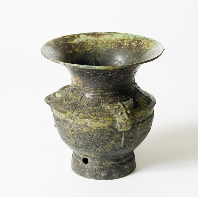 A BRONZE RITUAL WINE VESSEL, ZUN, SHANG /ZHOU DYNAST: The body and tall flared foot flat-cast with three large taotie masks, the taotie positioned below rams' heads cast in high relief on the shoulder between pairs of kui dragons, 8 7/8''H 8 3/8''Diam