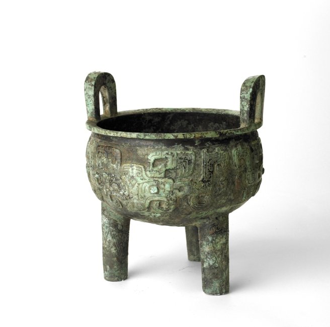 A BRONZE RITUAL TRIPOD VESSEL, DING SHANG DYNASTY: Raised on three columnar supports , the deep rounded sides cast in low relief between the legs with three taotie masks below a band of four pairs of bird-headed dragons, all reserved on a leiwen groun