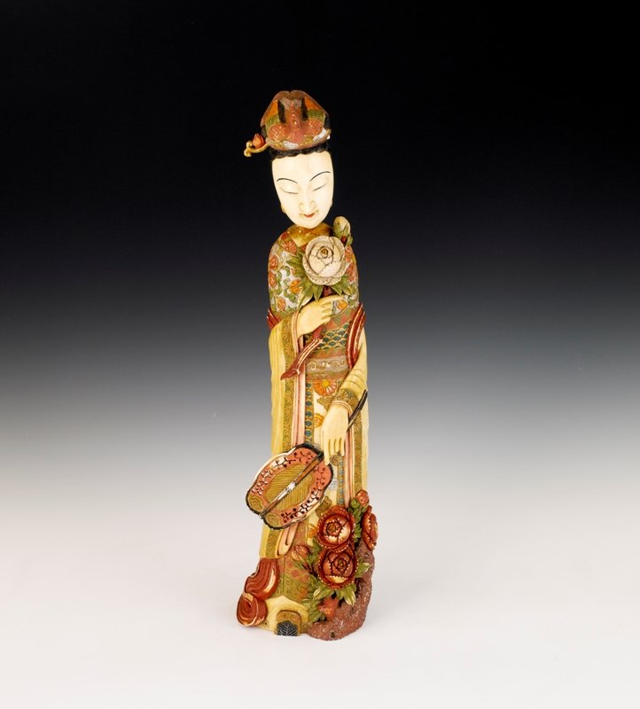 A colored ivory beauty, 19th Century: 19th Century Modeled wearing long robes, holding a spray of flower in her right hand and a traditional fan in her left hand, shown an age hairline crack on her face. H:14 3/4 in