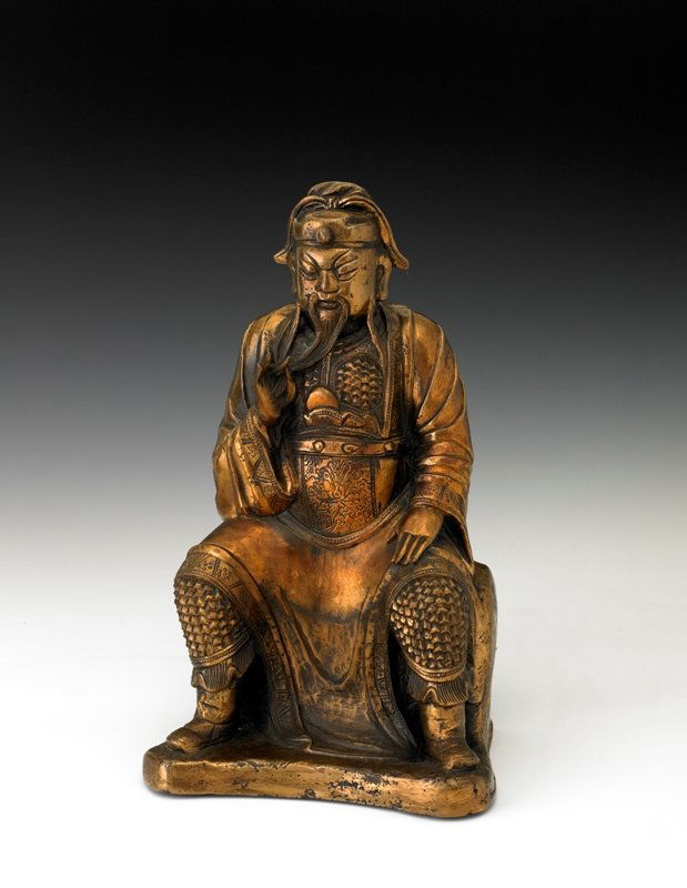 A gilt bronze figure of guandi, Ming dynasty: Ming dynasty The God of War seated on a stepped rockwork base with his left hand resting on his knee, his right raised to hold the end of his long beard, wearing a long robe, draped over one shoulder