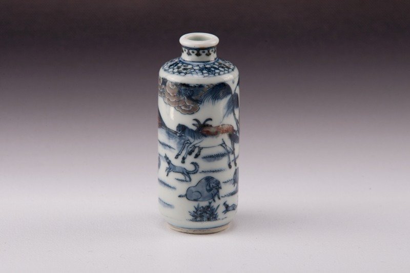 A blue-white -iron-red dragon snuff bottle, 19TH CE: 19TH CENTURY White/ blue porcelain color dress with numerous wild animals; mule, wolf, ram, ox and other mammals in a nature valley. 3 1/4 in. tall.