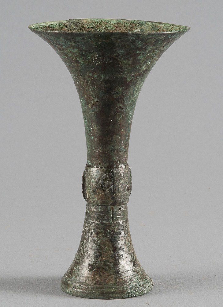 A BRONZE RITUAL WINE VESSEL, GU: WESTERN ZHOU DYNASTY The trumpet-shaped upper body is plain. The slightly convex mid-section is flat-cast with two taotie masks that are centered on a narrow flange and have conical pupils, and set be
