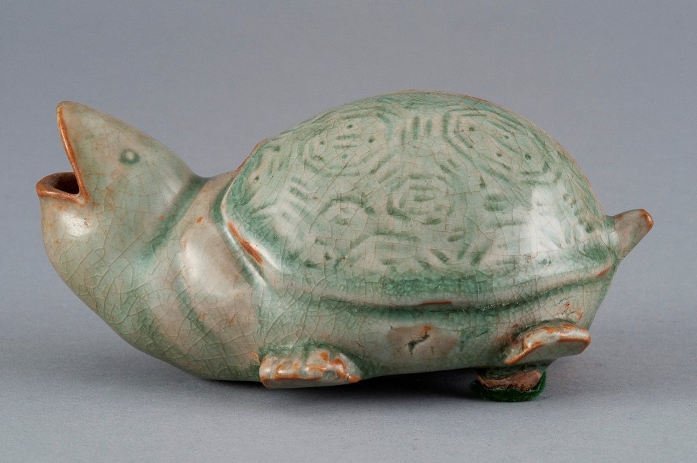 A CELADON-GLAZED TURTLE: MING DYNASTY The turtle is sea- green colour tone overall, all four legs grounded. The turtle's head is tilted up with its mouth open. The shell of the turtle has patterns of octogen- shapes. 6 1/16 i
